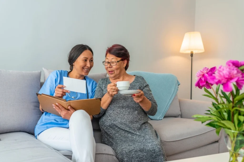 Happy nurse sitting with elderly client on couch in living room looking at photos together