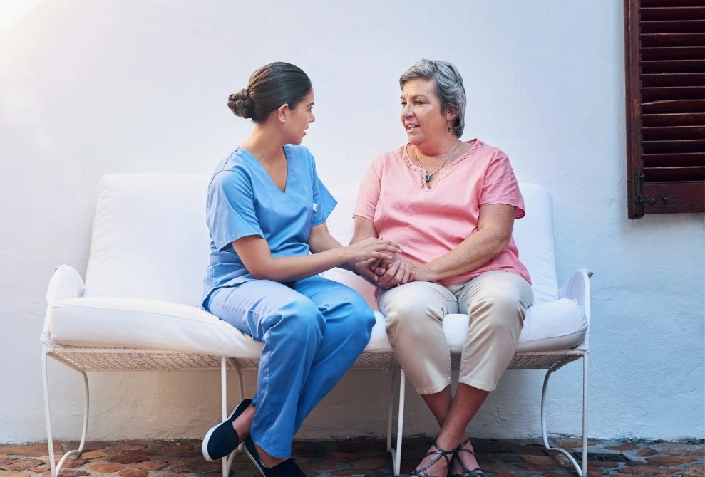 Casual nurse sitting with client on bench seat at home holding hands